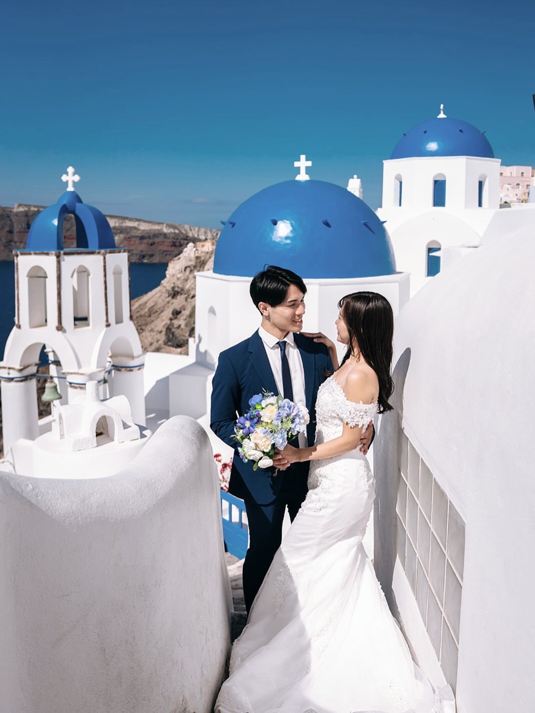Newlywed couple posing in wedding attire among whitewashed buildings and blue domes in Santorini.