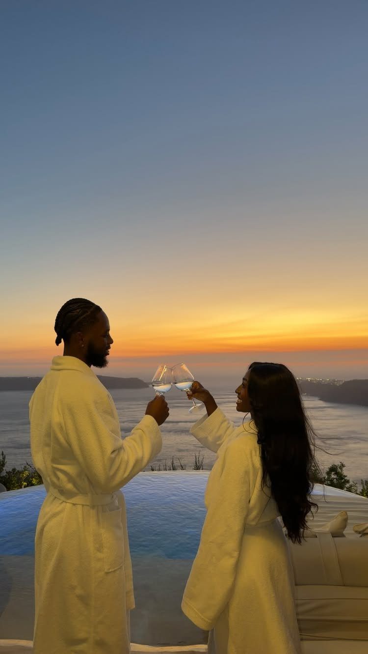 Couple in white bathrobes toasting with wine glasses by an infinity pool at sunset.