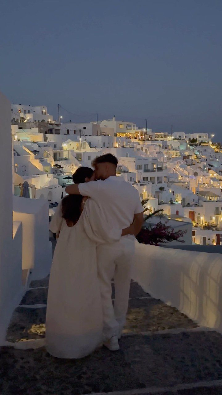 Couple in white embracing overlooking whitewashed buildings and lights in Santorini at twilight.