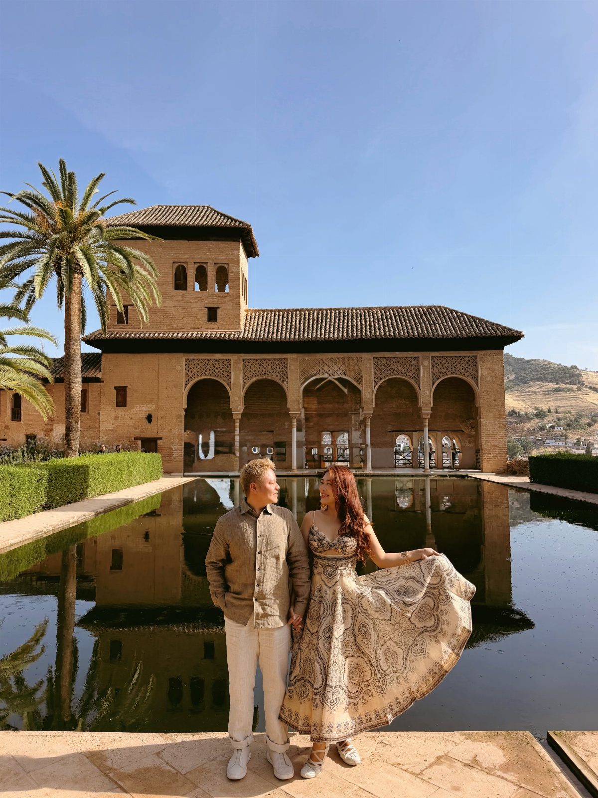 Couple posing by a reflecting pool in front of the Alhambra palace architecture.