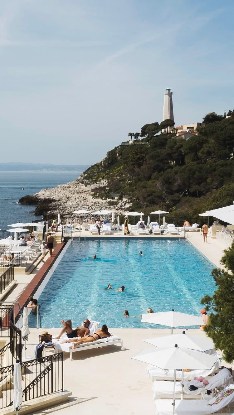 Luxury swimming pool with white umbrellas overlooking a rocky coast and white lighthouse.