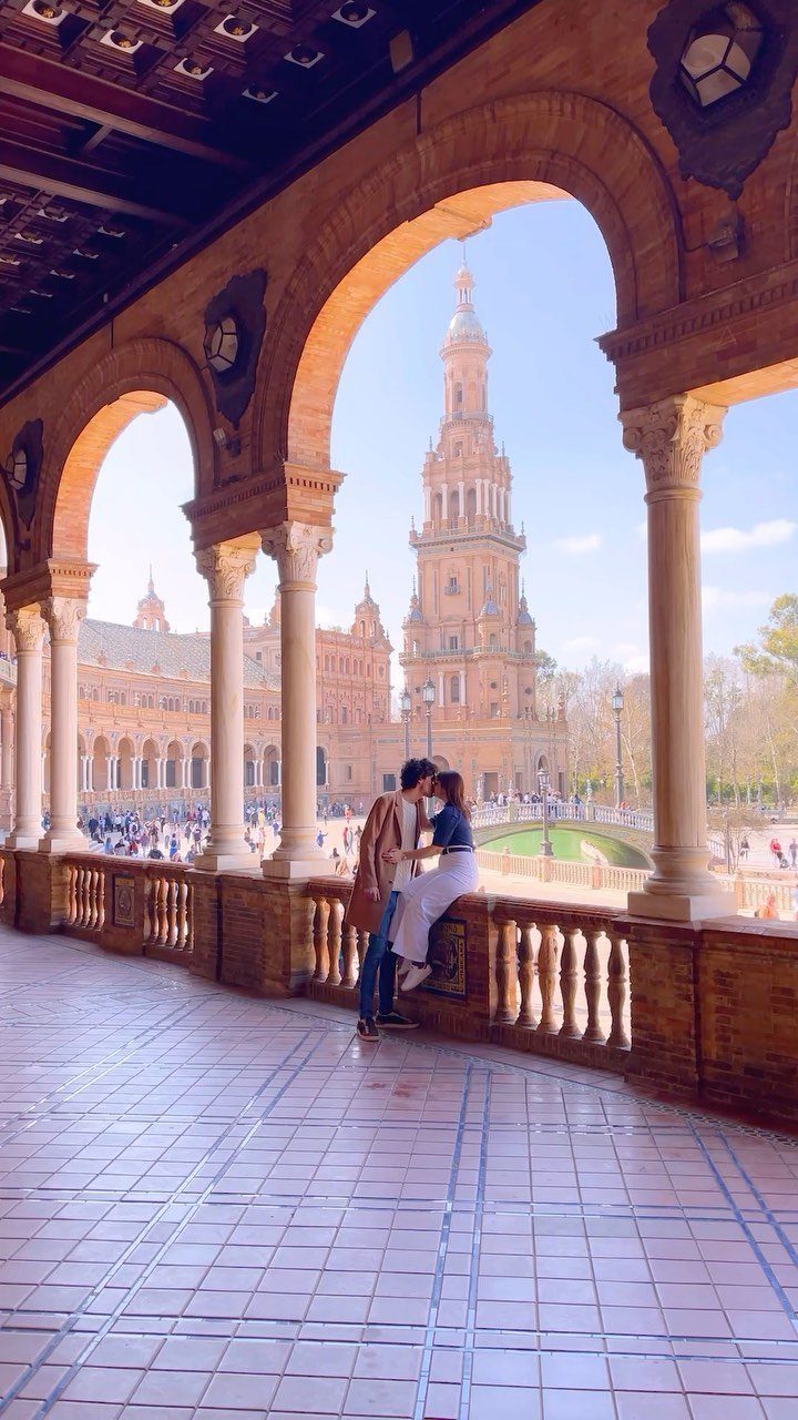 Couple embracing under stone arches with a tall Spanish tower in the background
