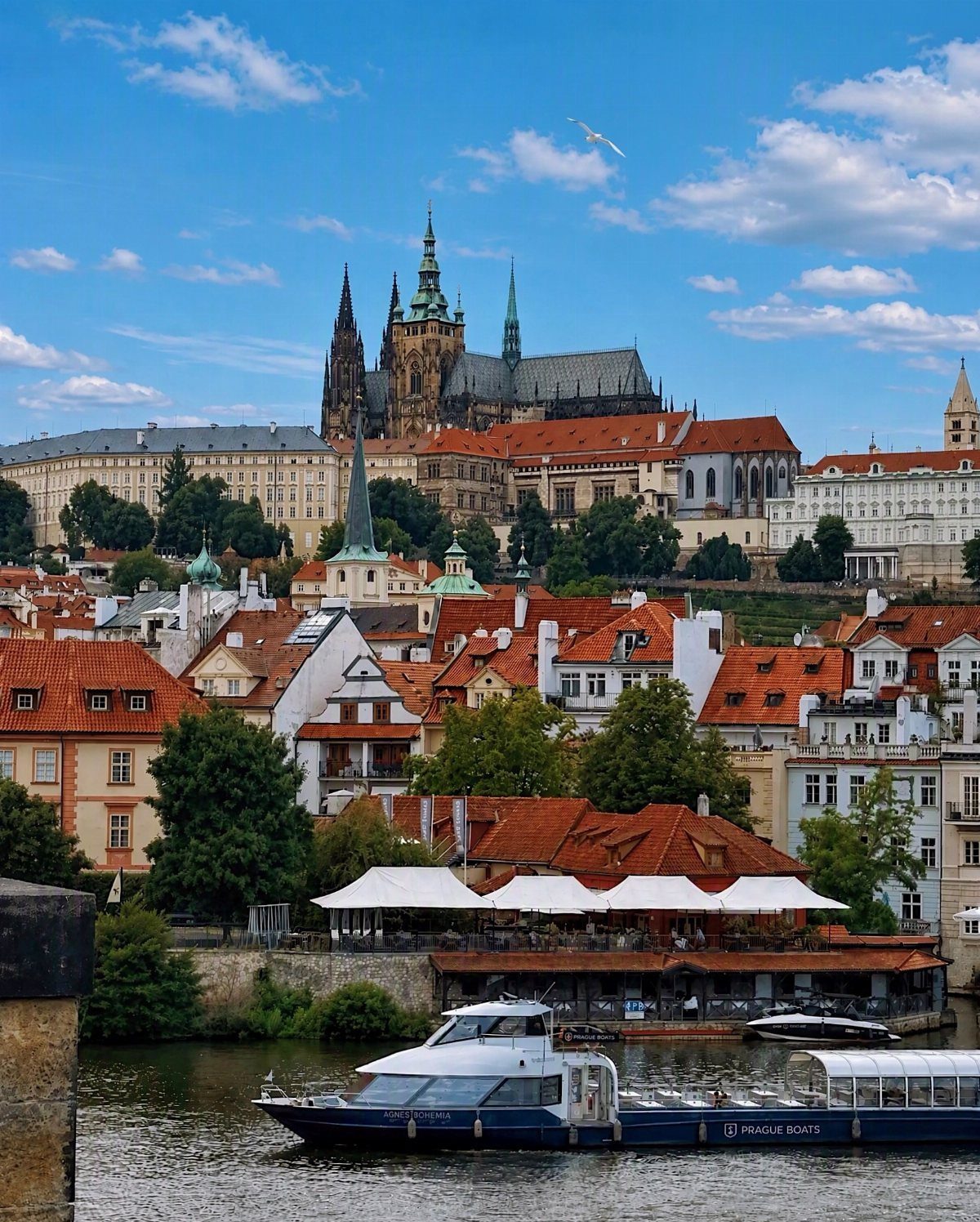 Prague cityscape with red roofs, a river boat, and St. Vitus Cathedral