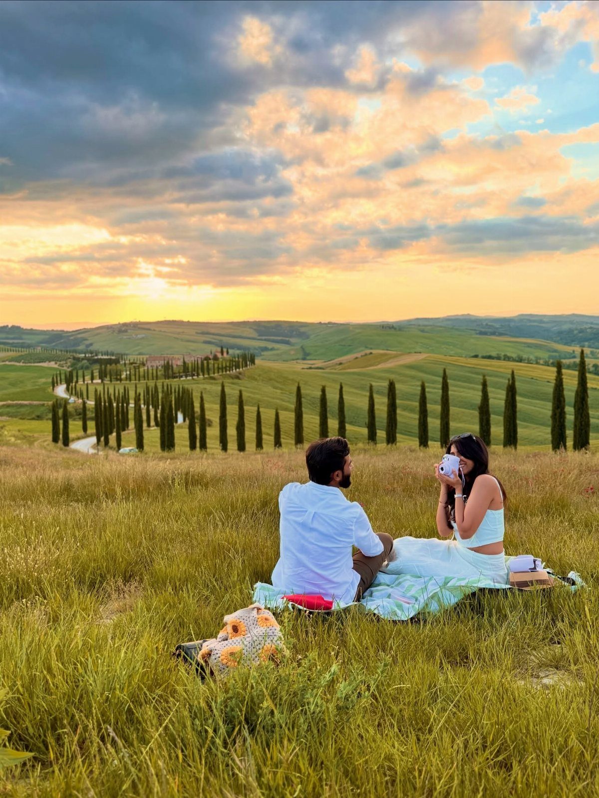 Couple sitting in a grassy field overlooking rolling hills and cypress trees at sunset.