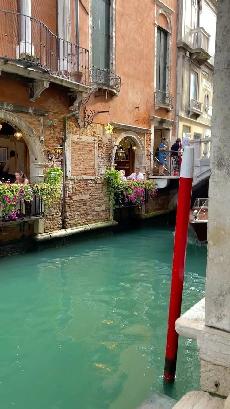 Turquoise canal in Venice with orange buildings, flower boxes, and a red mooring pole.