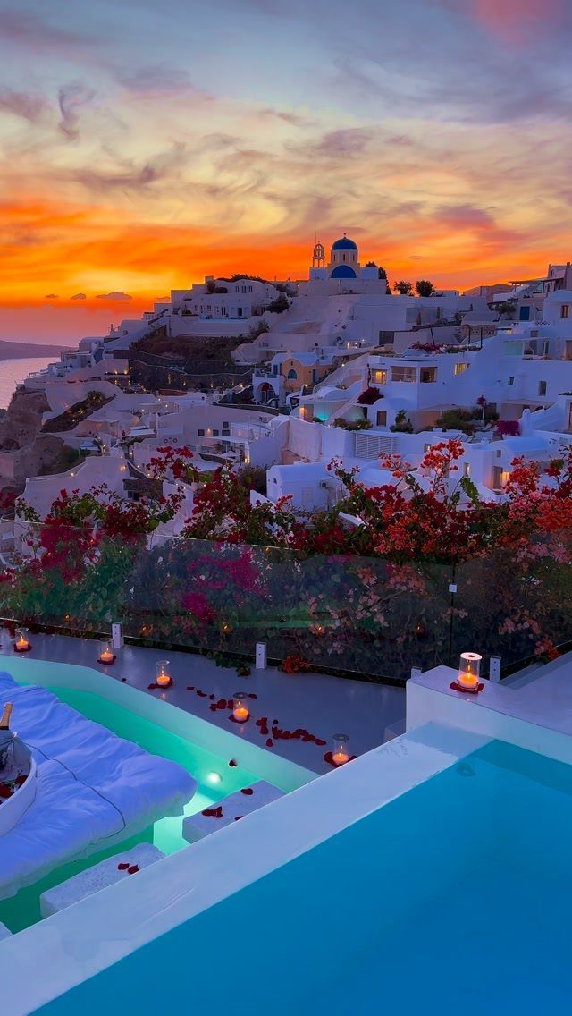 Infinity pool with rose petals overlooking whitewashed buildings and blue domes at sunset.