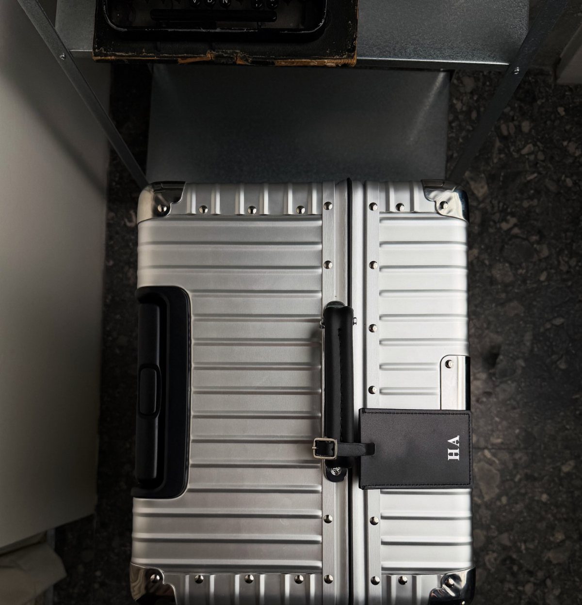 Silver aluminum suitcase with a black leather luggage tag on a stone floor