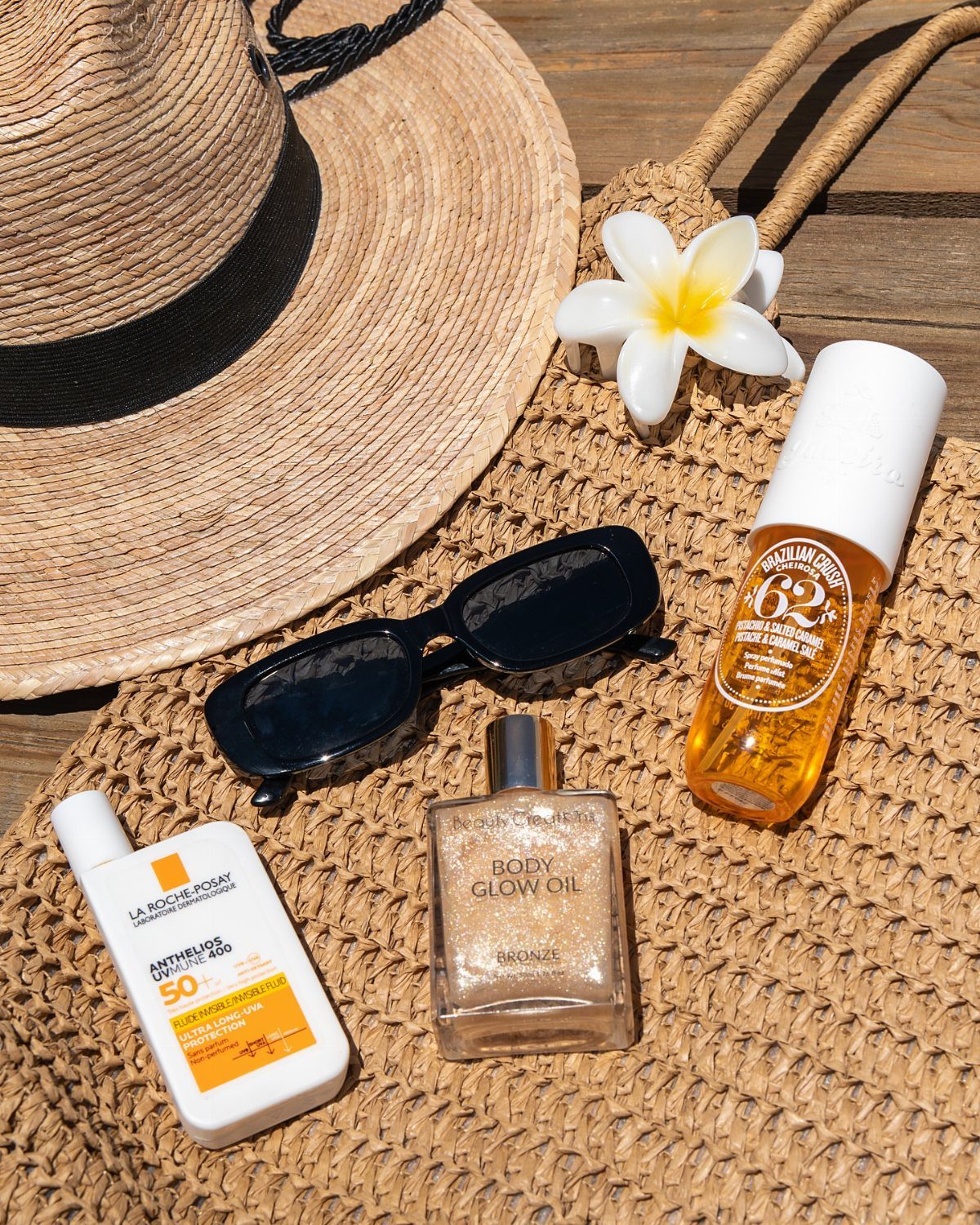 Sunscreen, body oil, sunglasses, and a straw hat arranged on a woven bag.