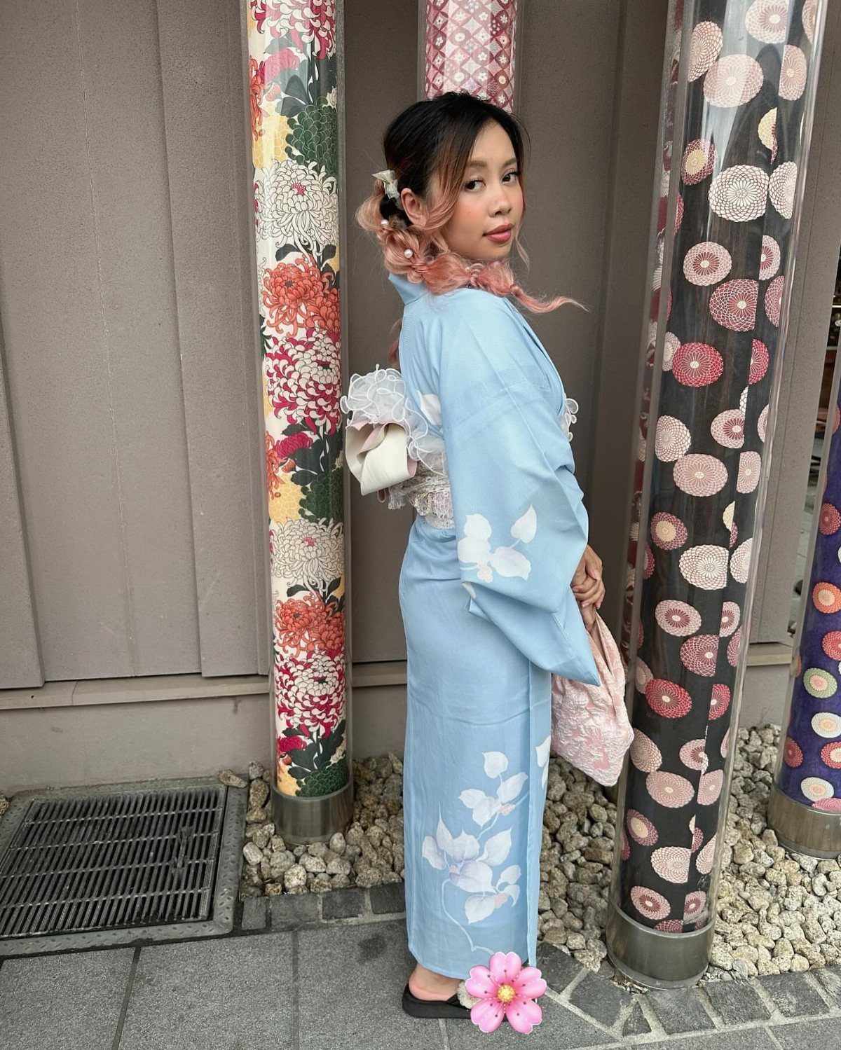Woman in a light blue floral kimono standing by decorative floral pillars outdoors.
