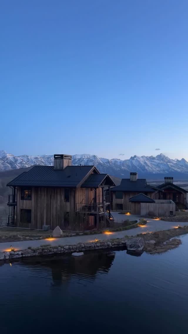 Wooden lodge cabins by a lake with snow-capped mountains under a twilight sky.