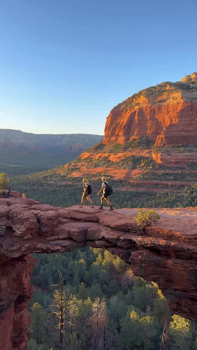 Two hikers crossing a red rock bridge with towering red sandstone cliffs behind them.