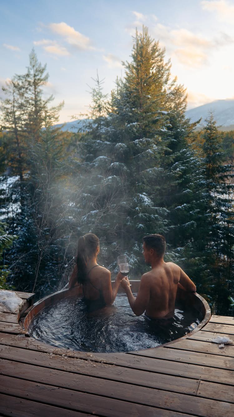 Couple soaking in a steaming outdoor hot tub surrounded by snowy evergreen trees.