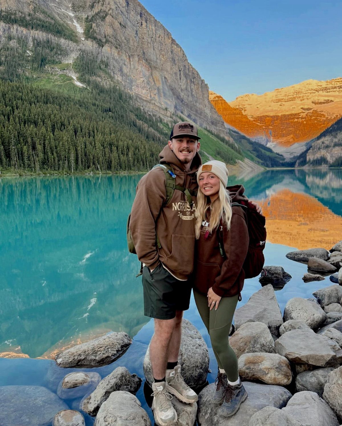 Couple in hiking clothes posing by a turquoise lake with rugged mountains behind them.