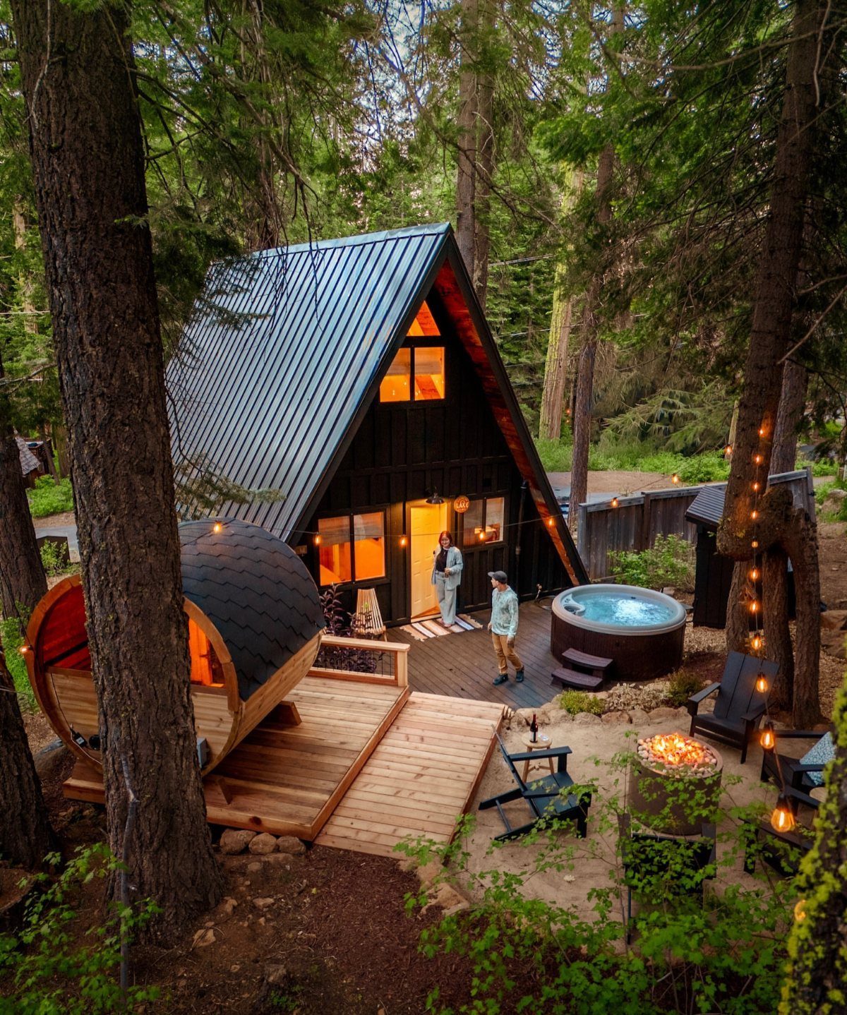 Black A-frame cabin in a forest with a hot tub and barrel sauna.