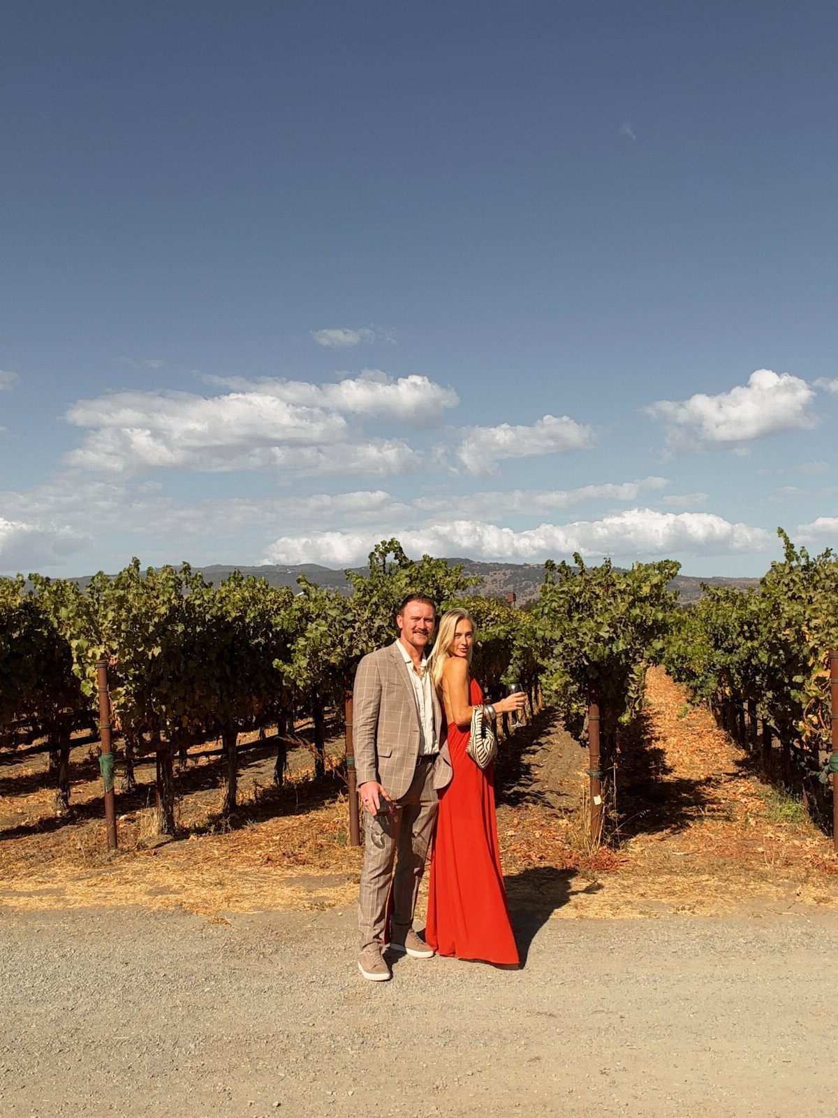 Couple in formal attire posing in a vineyard with rows of grapevines.