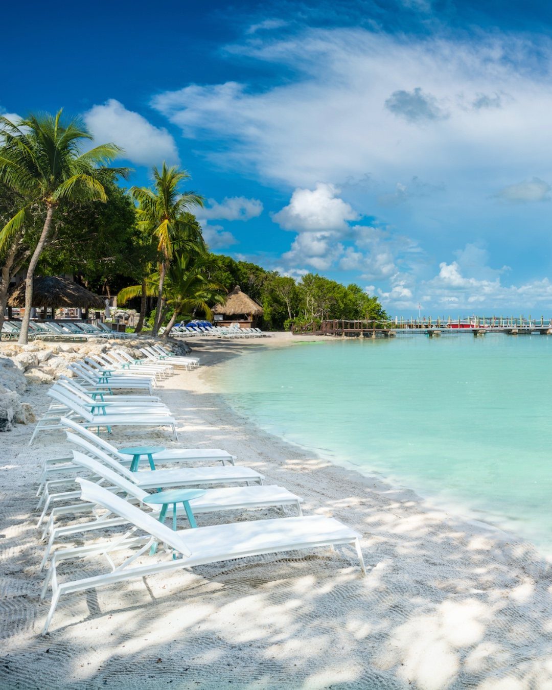 White lounge chairs on a sandy beach with turquoise water and palm trees.