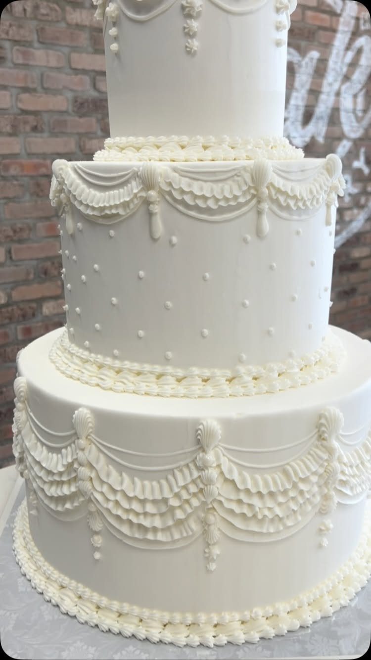 Three-tier white wedding cake with ornate frosting swags and pearls against brick wall