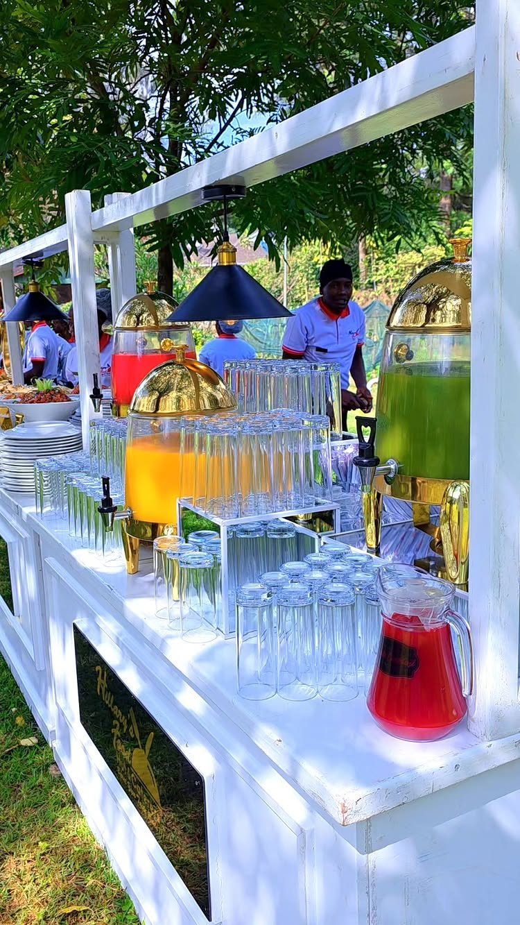 Outdoor catering station with gold beverage dispensers and rows of clear glasses.