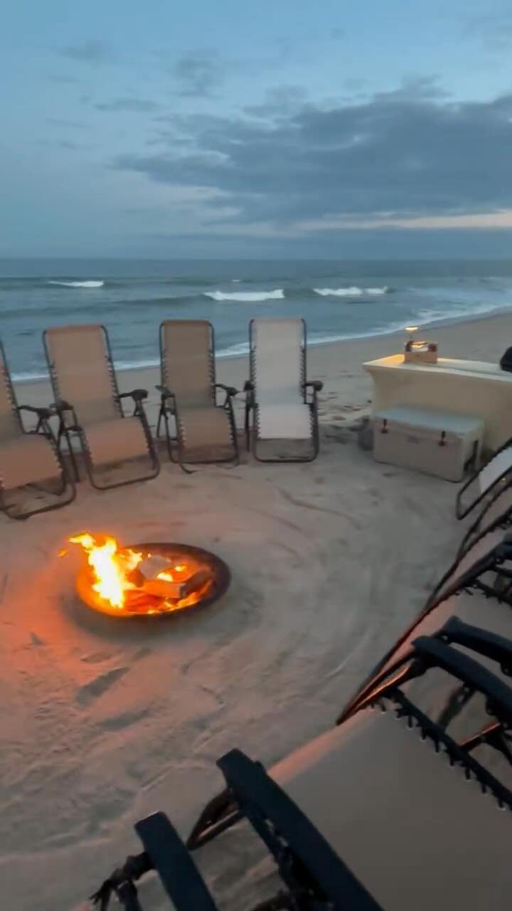 Beach fire pit with lounge chairs on the sand overlooking the ocean at dusk.