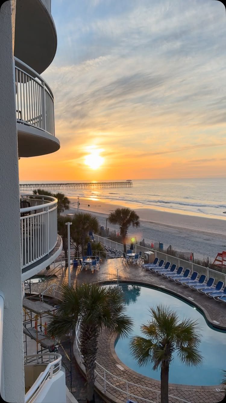 Resort balcony view of a swimming pool, beach, and pier at sunset.