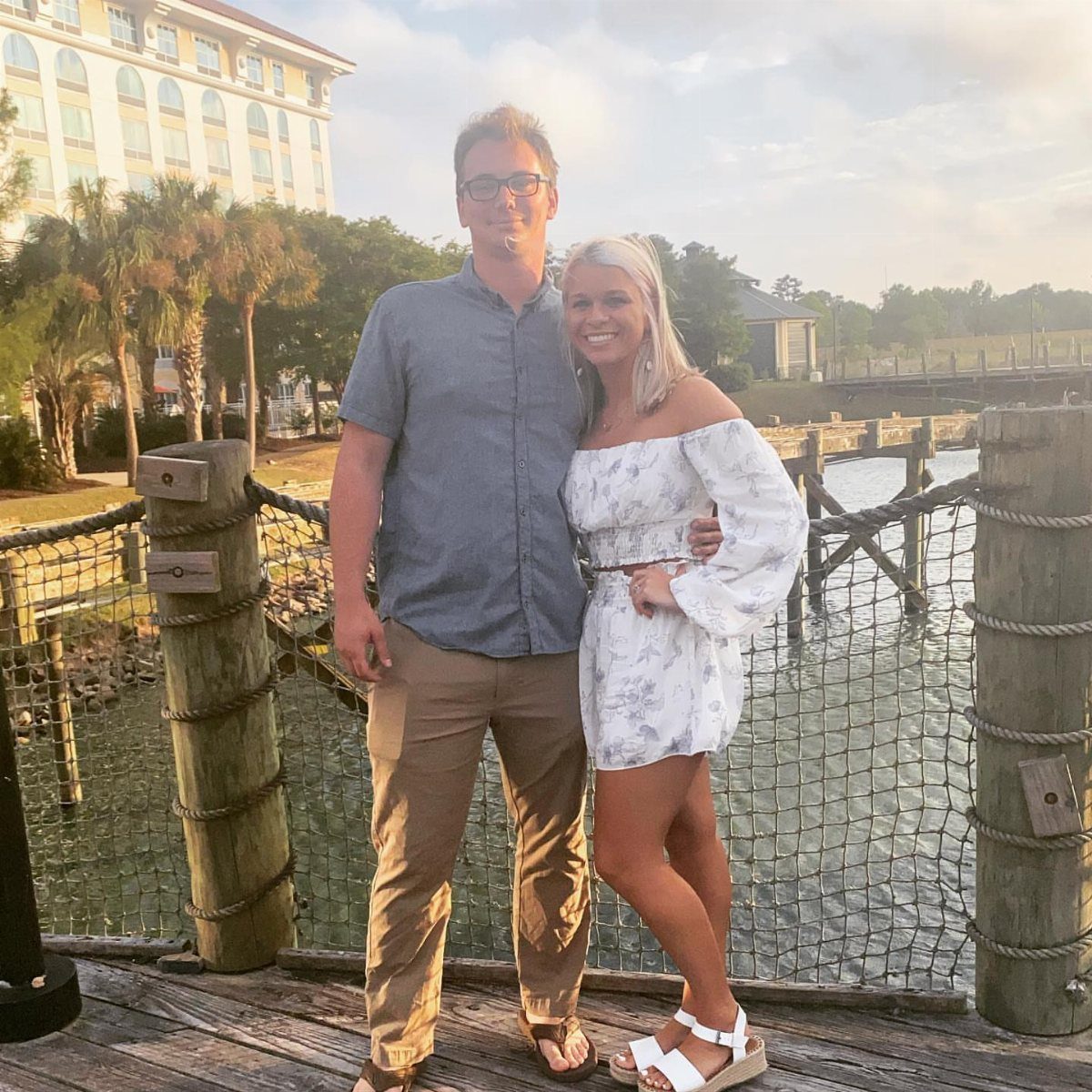 Smiling couple posing on a wooden pier with a hotel and palm trees behind.