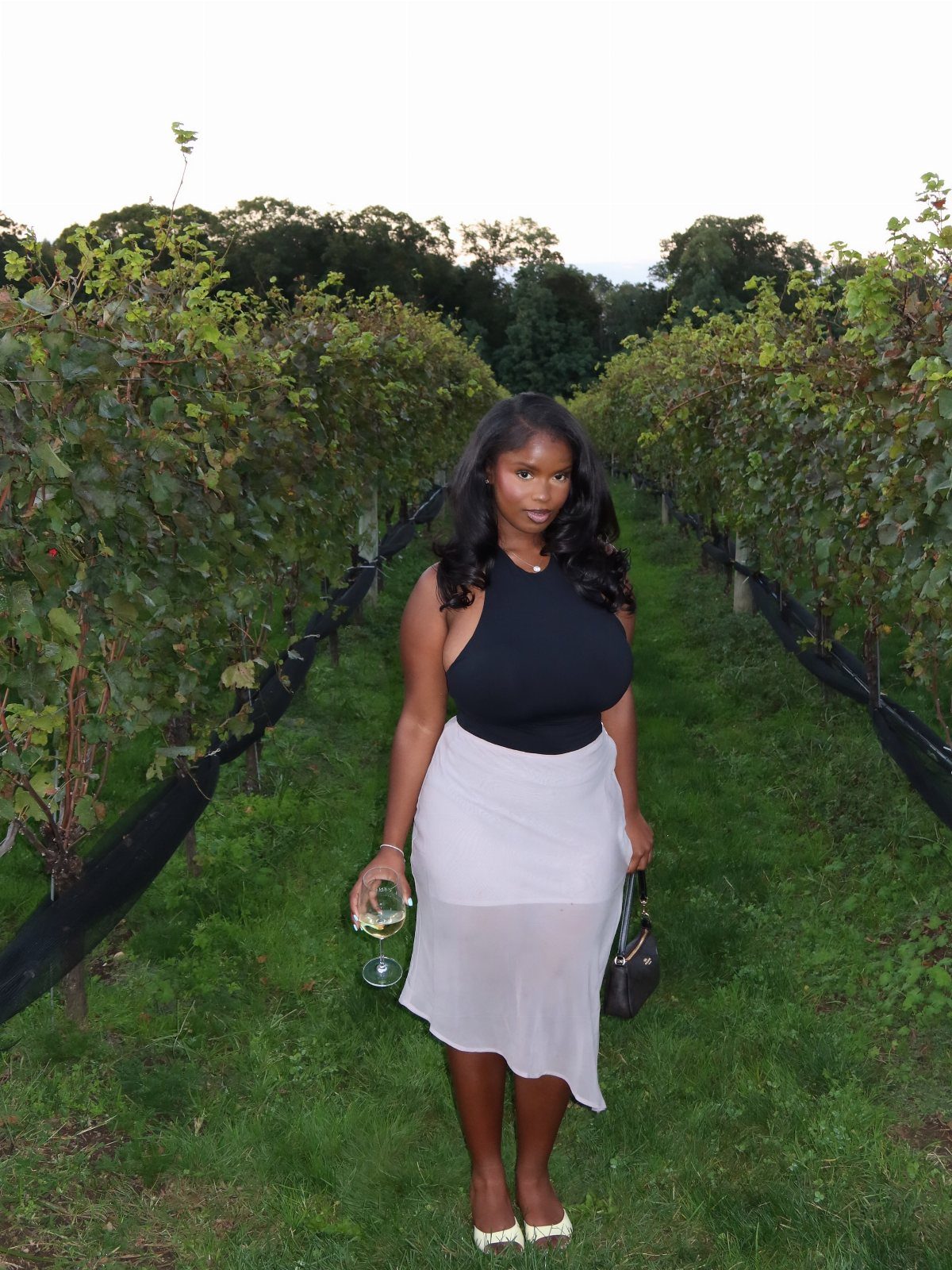 Woman holding a wine glass while standing between rows of grapevines in a vineyard.