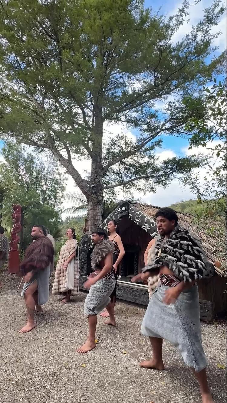 Maori people in traditional woven clothing standing before a carved wooden building.