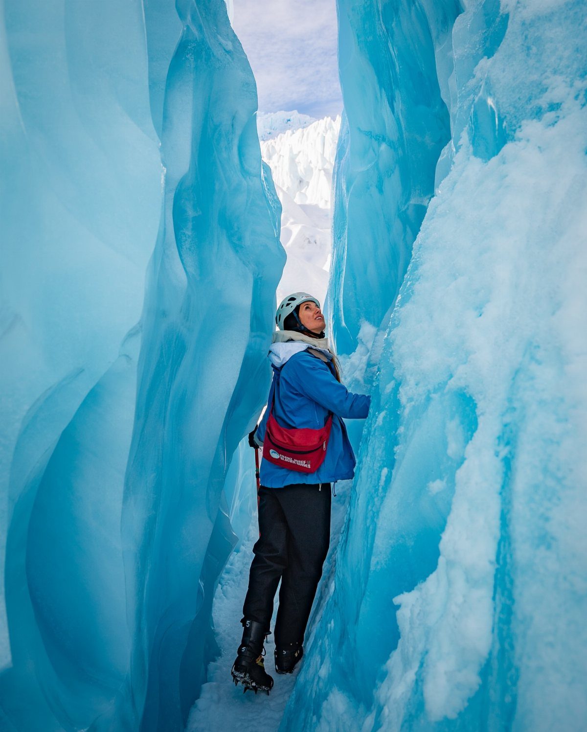Person in hiking gear standing between towering walls of blue glacial ice.