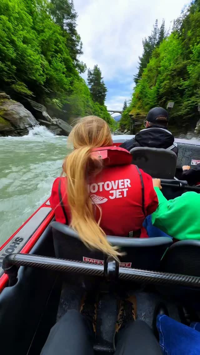 Woman in red life jacket riding a jet boat through a forested river canyon.