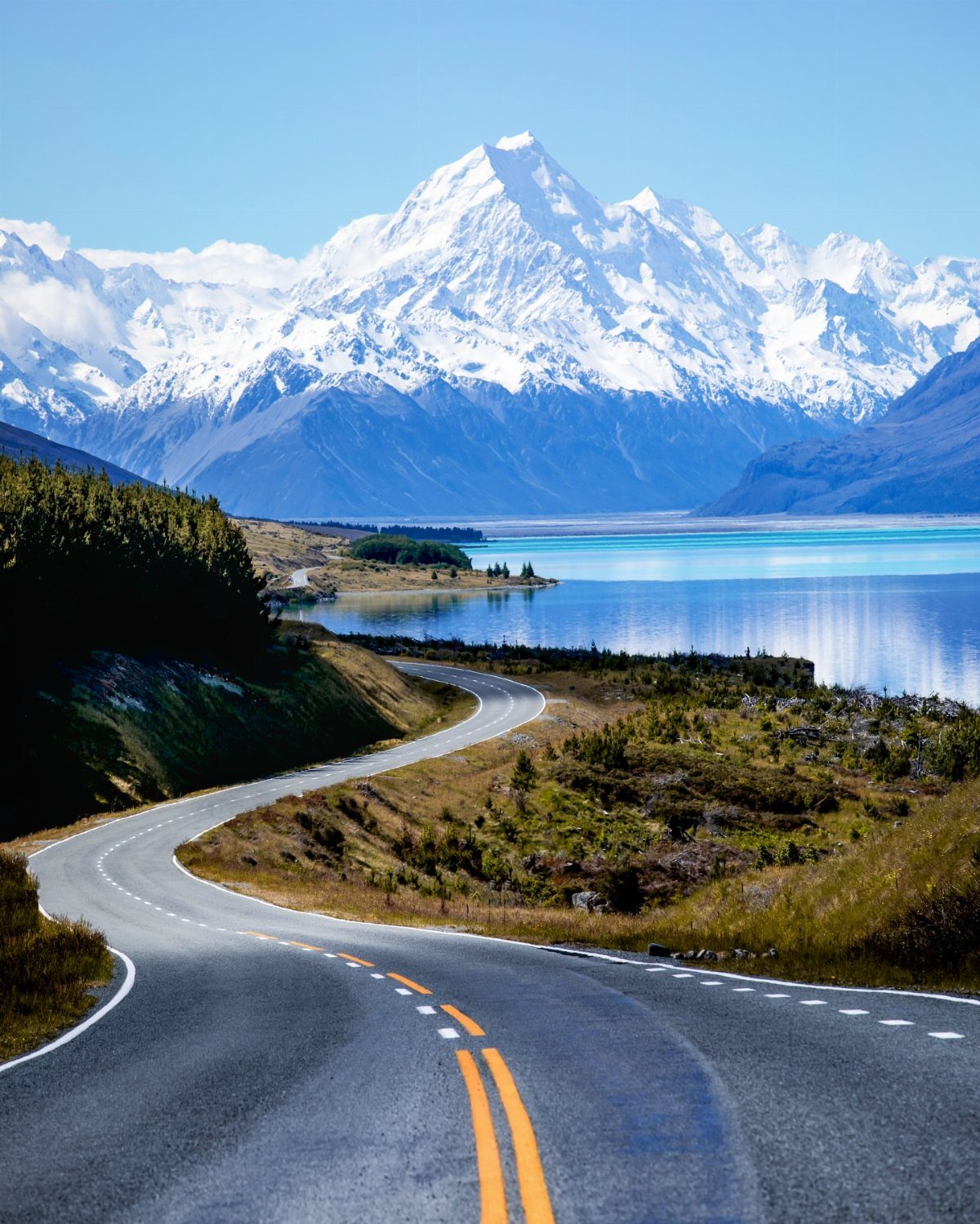 Winding road leading toward a turquoise lake and snow-capped mountains in New Zealand.