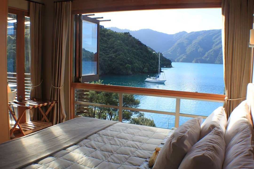 Bedroom window overlooking a turquoise bay with a sailboat and forested mountains.