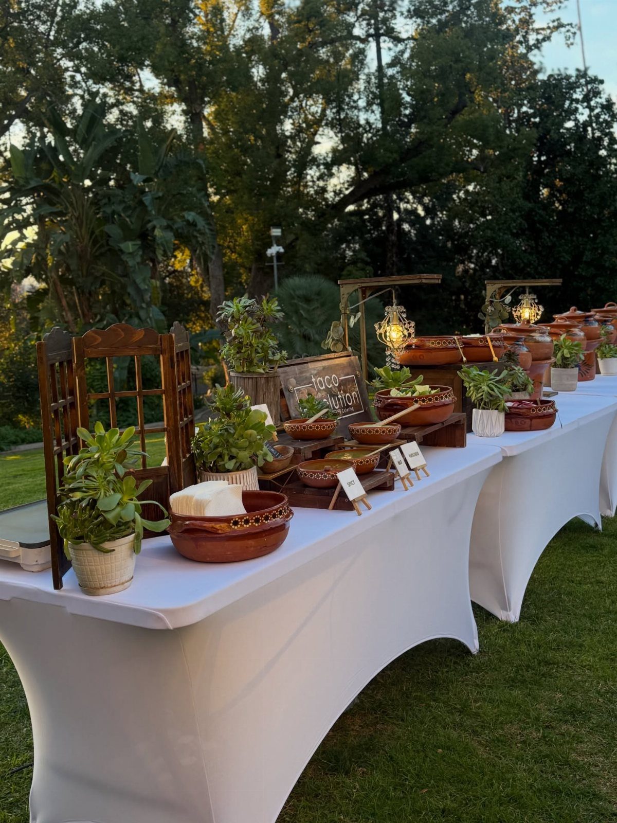 Outdoor food station with clay bowls and plants on white draped tables.