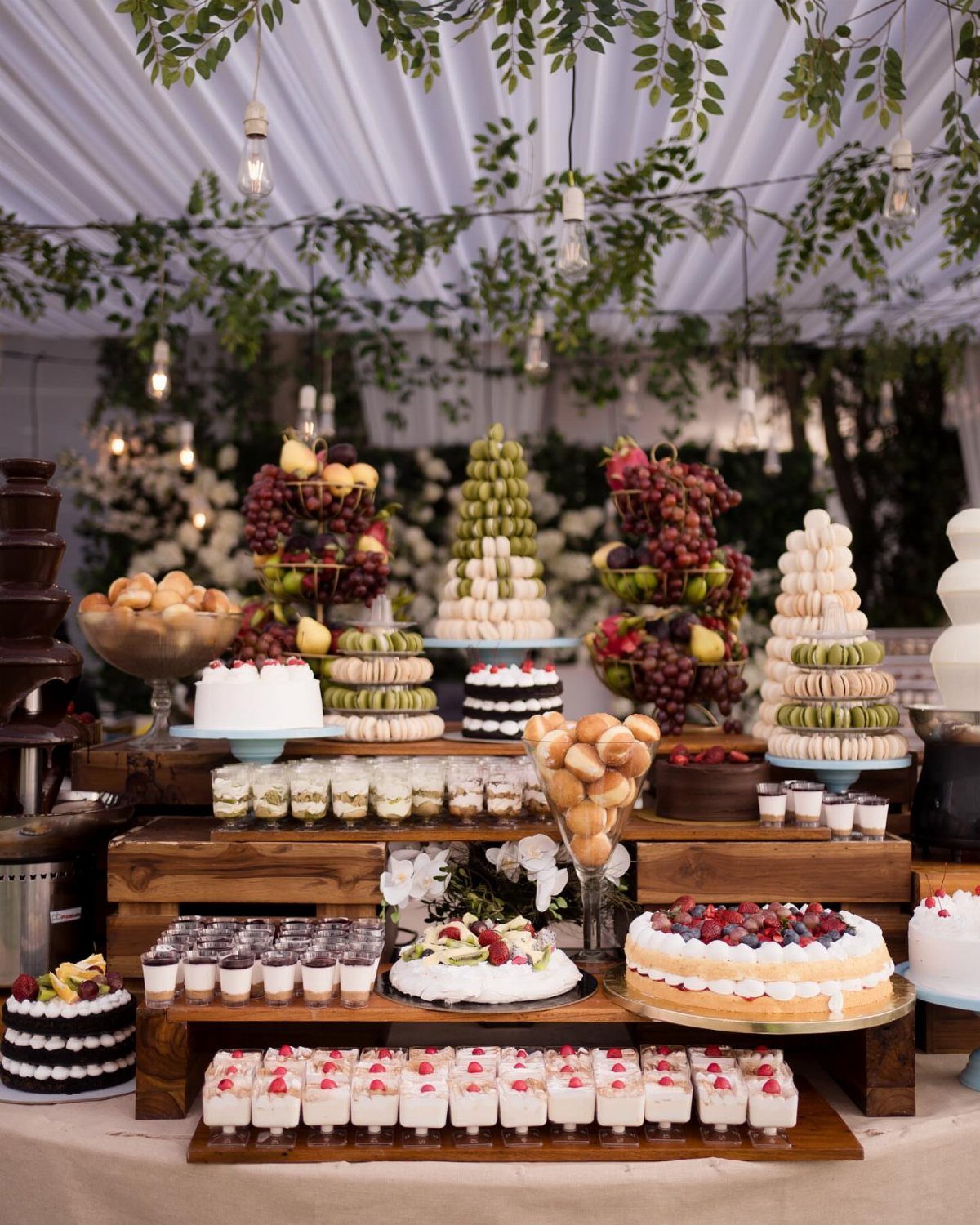 Lavish dessert table with cakes, macarons, and fruit towers under a white tent.