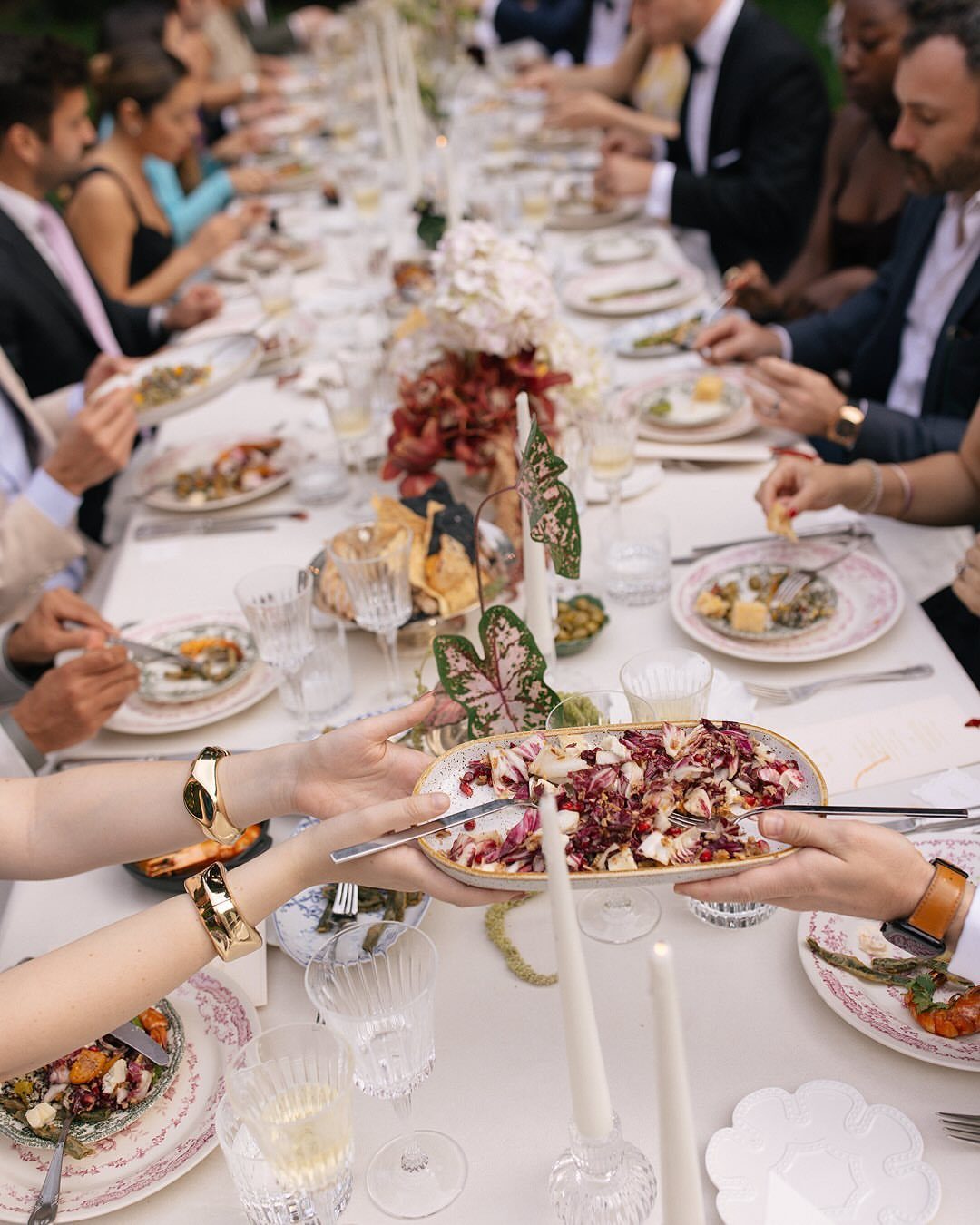 Guests passing a platter of appetizers across a long, decorated outdoor wedding table.