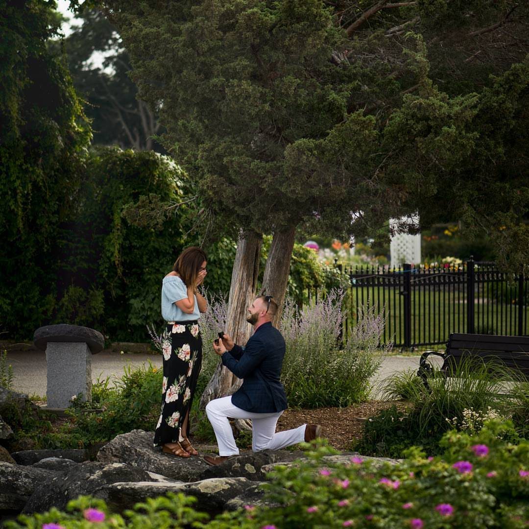 Man kneeling on a rocky garden path proposing to a woman in floral skirt