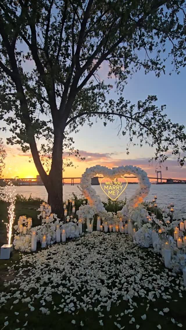 Heart-shaped floral arch with proposal sign on white petal path at sunset