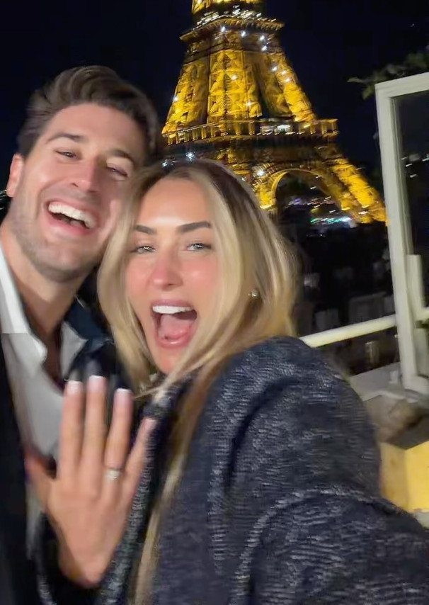 Smiling couple posing with an engagement ring in front of the Eiffel Tower.