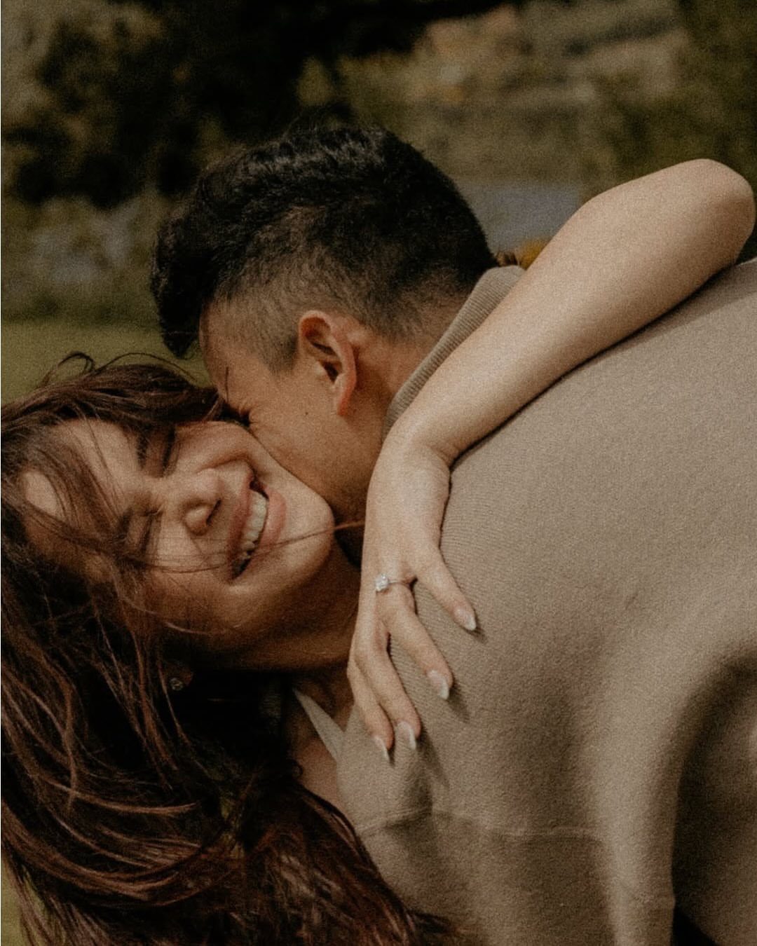 Smiling woman being embraced and kissed by a man, showing an engagement ring.