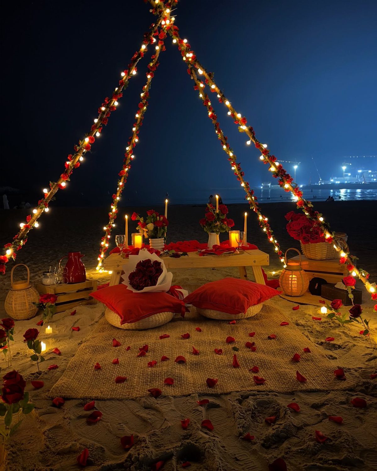 Romantic beach picnic setup with red cushions, rose petals, and fairy lights at night.