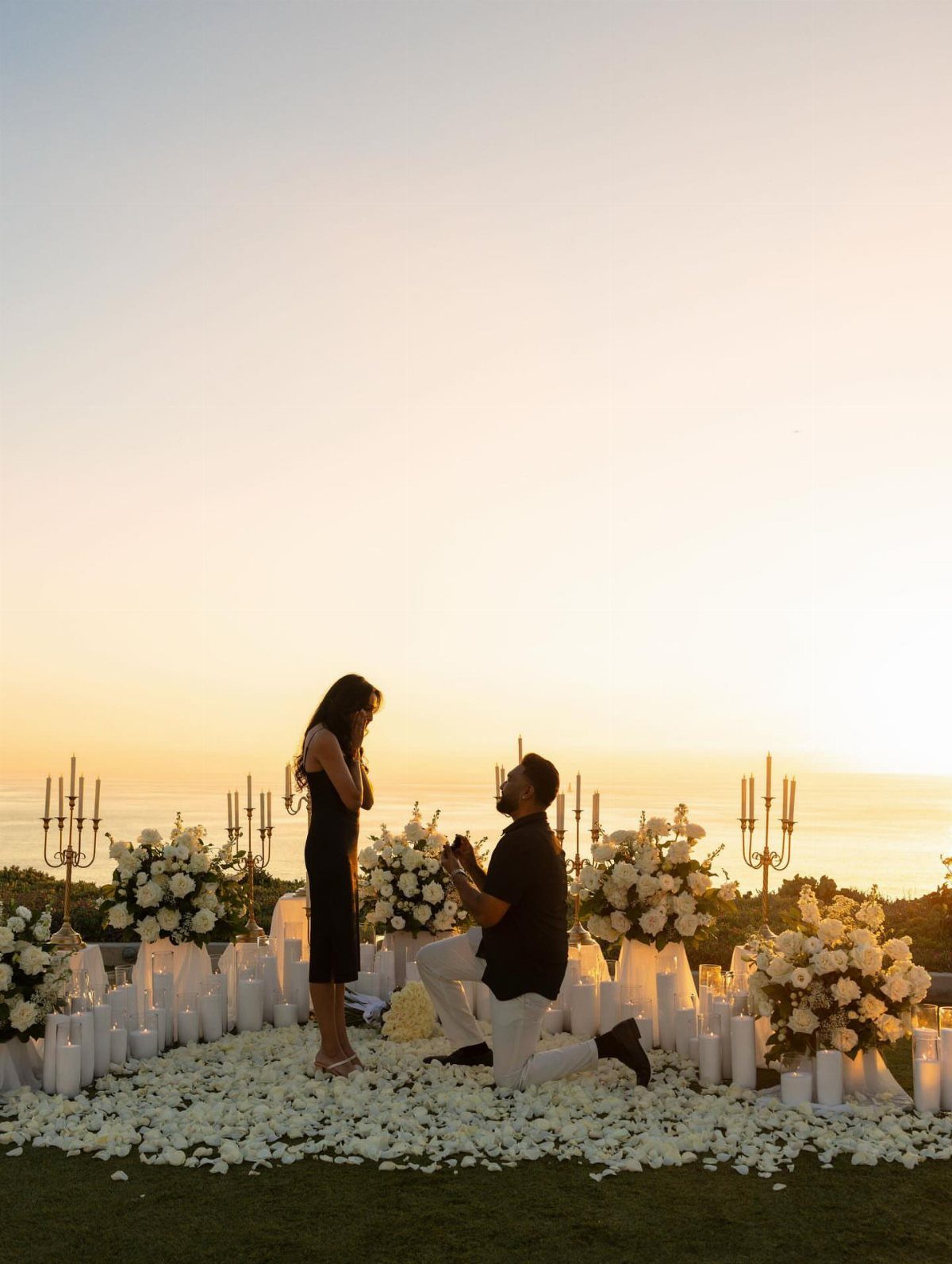 Man kneeling to propose to a woman amidst white roses and candles at sunset.