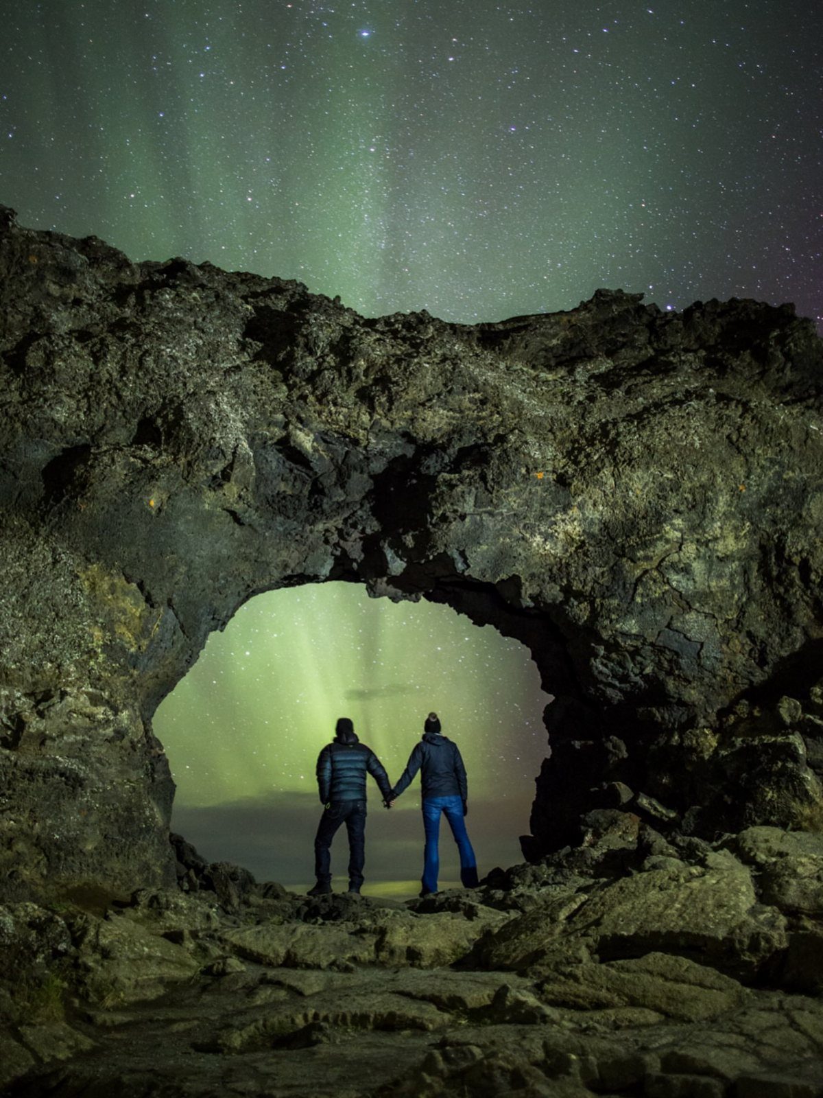 Couple holding hands under a rock arch beneath the green northern lights.