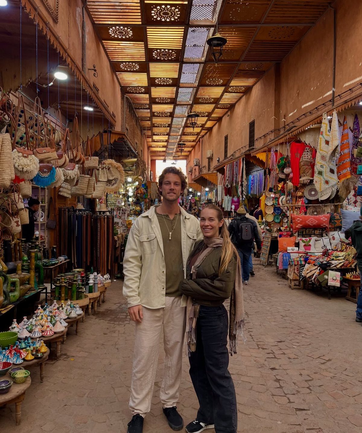 Couple smiling in a vibrant Marrakech market with colorful textiles and ceramics.