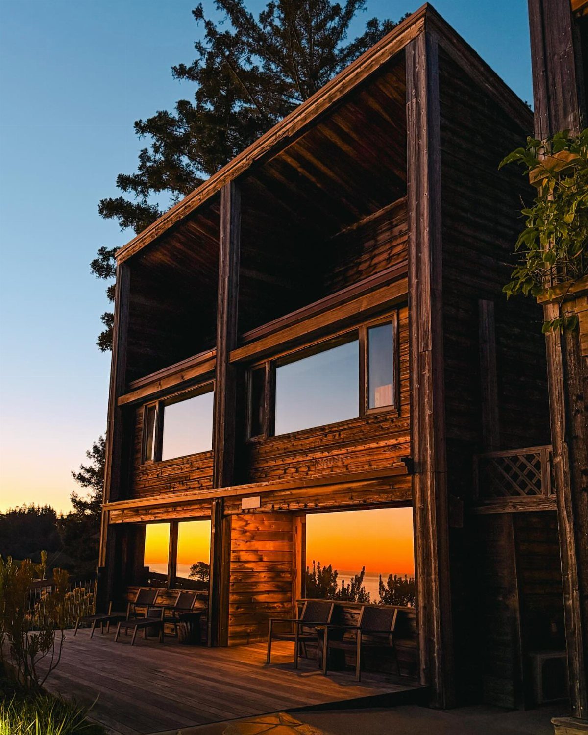 Wooden cabin with large windows reflecting a sunset, surrounded by tall evergreen trees.