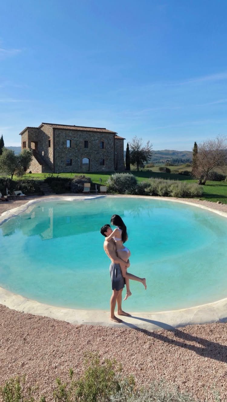 Couple hugging by a turquoise pool with a stone villa and rolling hills.