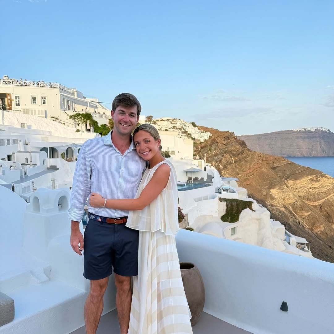 Smiling couple posing on a white terrace overlooking Santorini's white-washed buildings and cliffs.