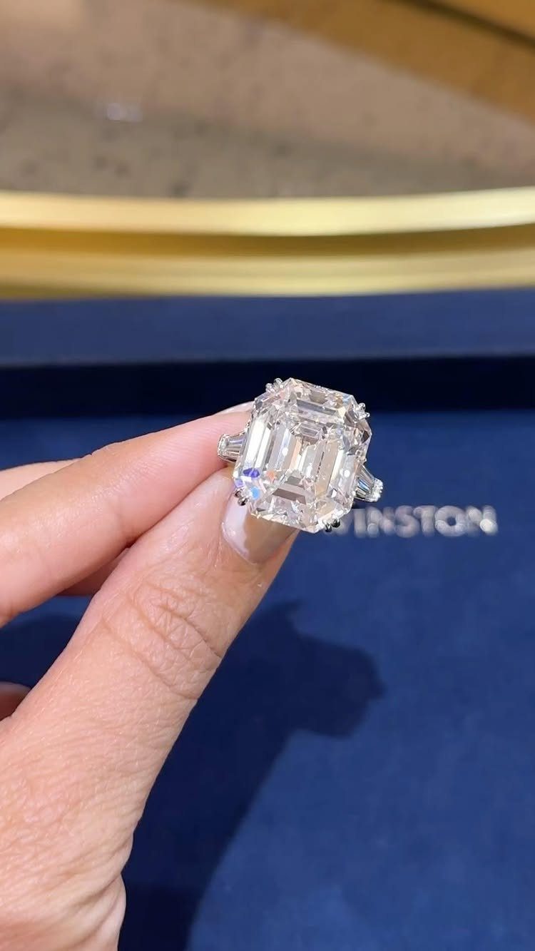 Emerald-cut diamond engagement ring held by a hand against blue velvet background.
