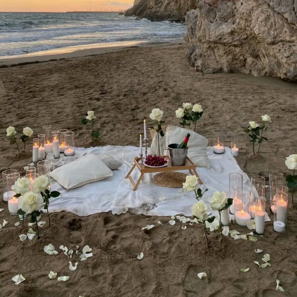 Romantic beach picnic setup with white roses, candles, and champagne on a blanket.