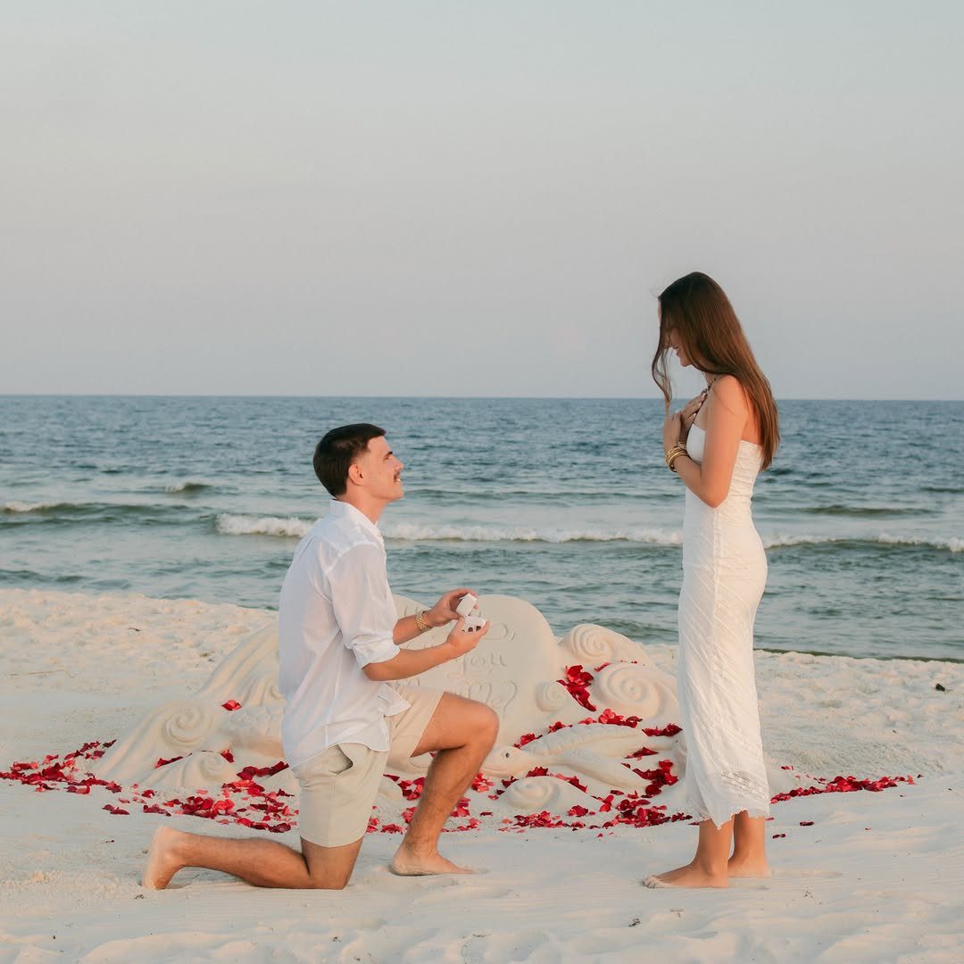 Man proposing to a woman on a beach next to a large sand sculpture.