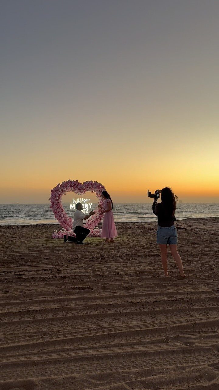 Man proposing to a woman on a beach with a floral heart backdrop.