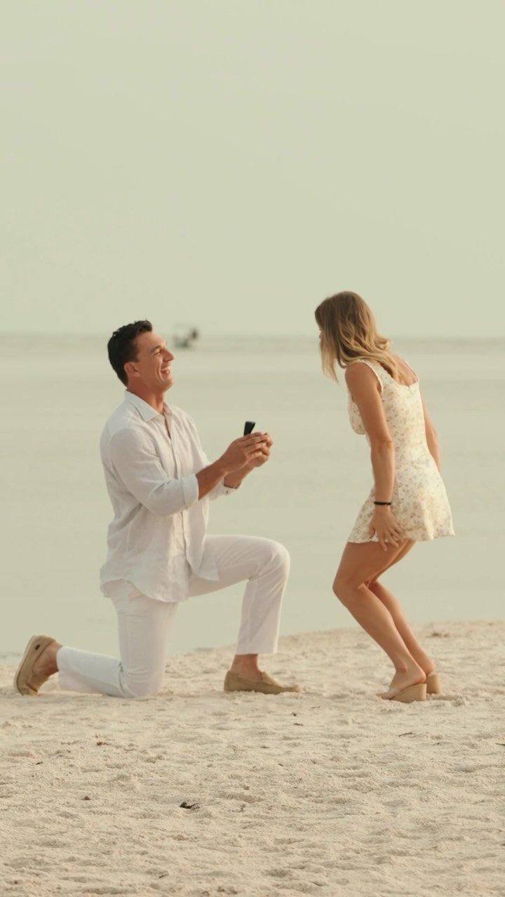 Man kneeling on a sandy beach proposing to a woman in a white dress.